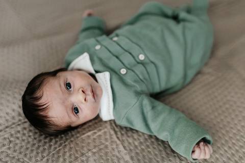 Bebê recém-nascido de roupa verde deitado de costas na manta e olhando para a câmera.'