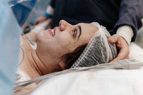 Mãe deitada no centro cirúrgico sorrindo aliviada durante o procedimento do parto.'