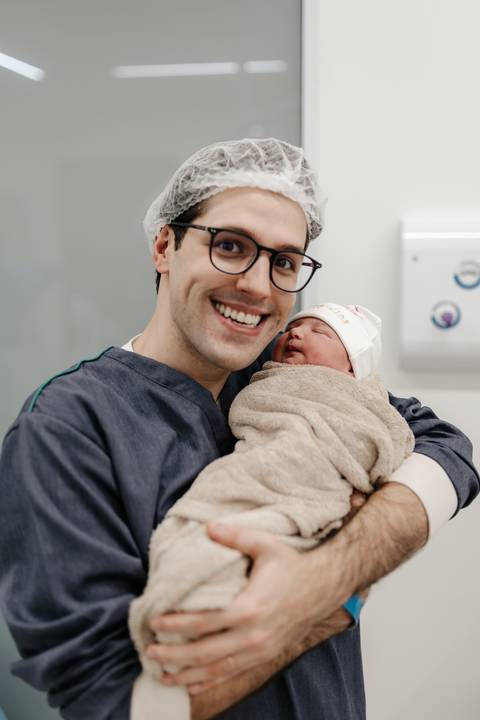 Pai sorridente vestindo pijama cirúrgico e segurando o bebê recém-nascido no colo.'