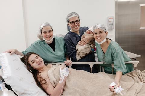 Retrato da família no centro cirúrgico com a equipe médica ao redor da mãe e bebê.'