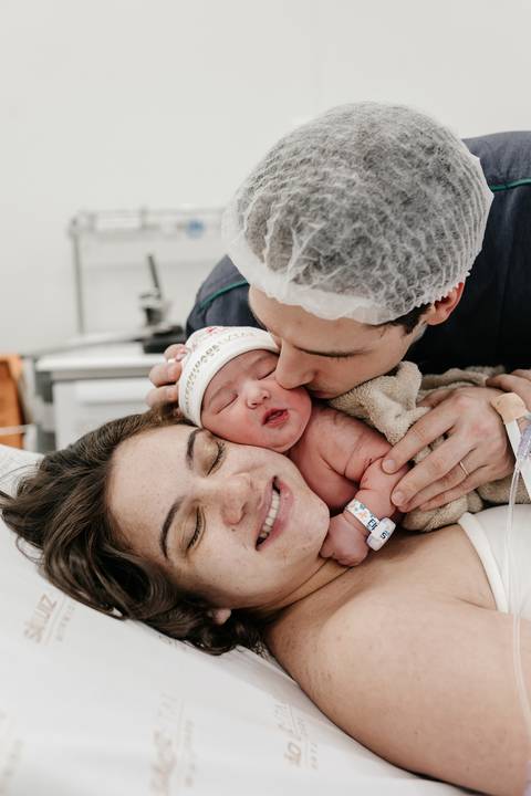 Pai dando um beijo carinhoso na bochecha do bebê enquanto a mãe sorri deitada.'
