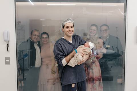 Pai segurando o recém-nascido no colo e acenando para a família através do vidro da sala.'