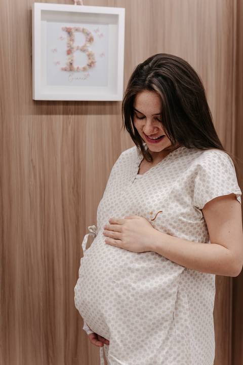 Plano médio da gestante sorrindo e olhando para a barriga com as mãos apoiadas sobre ela.'