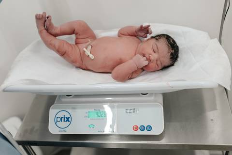 Bebê recém-nascido sendo pesado na balança pediátrica digital no berçário hospitalar.'