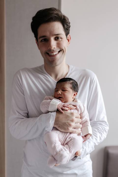 Pai sorridente vestindo blusa branca segurando o bebê no colo no quarto do hospital.'