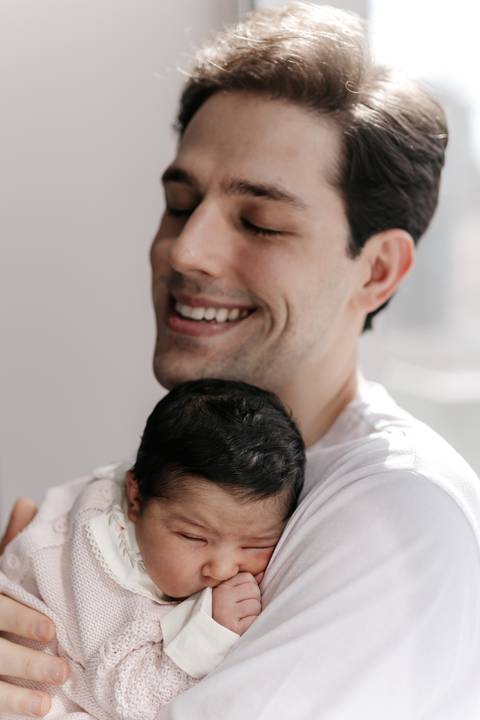 Retrato aproximado do pai sorrindo com o bebê aconchegado junto ao seu peito.'