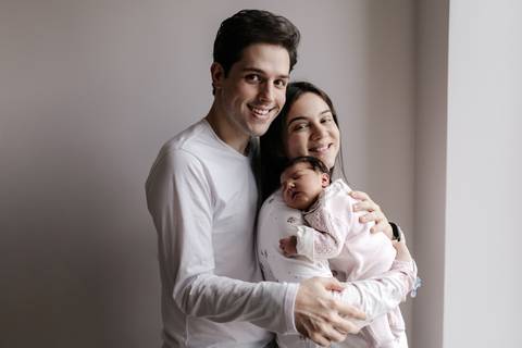 Retrato do casal sorridente abraçado com o bebê no colo no quarto da maternidade.'