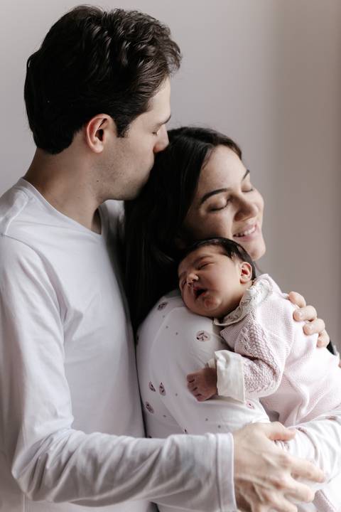 Pai e mãe de olhos fechados encostados no bebê adormecido em um momento de carinho.'