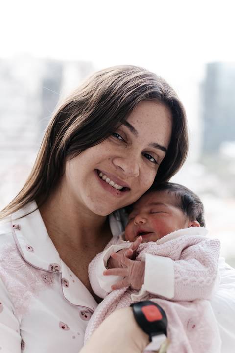 Mãe sorridente em close-up segurando o bebê adormecido encostado em seu rosto.'