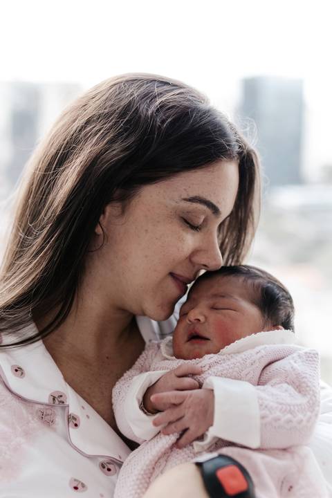 Mãe de olhos fechados dando um beijo carinhoso na testa do bebê dormindo.'