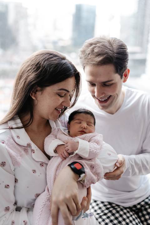 Casal sorrindo e olhando encantado para o bebê no colo da mãe no quarto da maternidade.'