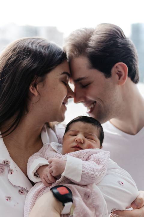 Retrato do casal de rostos colados e sorrindo enquanto seguram o bebê dormindo.'