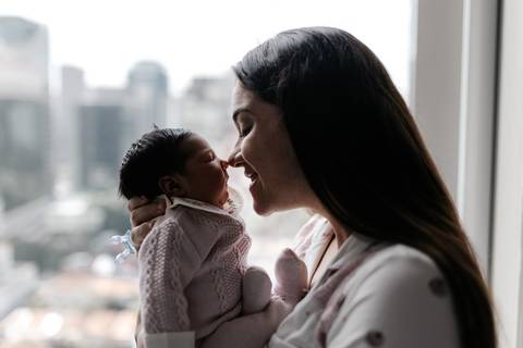 Silhueta da mãe e do bebê em frente à janela do quarto com vista da cidade ao fundo.'