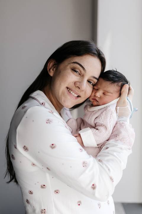 Mãe sorrindo para a câmera enquanto segura o bebê aconchegado em seus braços.'