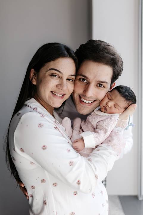 Retrato da família reunida com pai e mãe sorrindo radiantes com o bebê dormindo.'