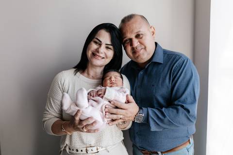 Retrato dos avós maternos sorrindo ao lado da neta recém-nascida na maternidade.'