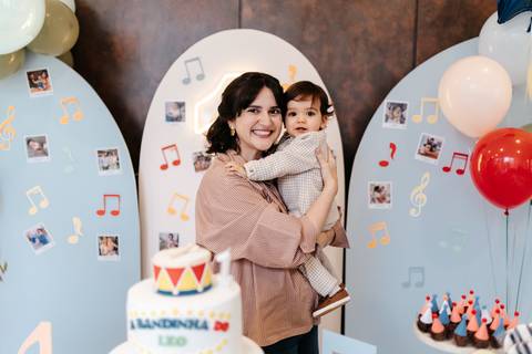Mãe sorridente segurando o aniversariante no colo em frente ao painel decorativo musical.'