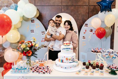 Pais e o bebê posando juntos atrás da mesa do bolo decorada com balões e doces.'