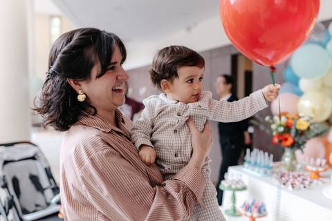 Mãe segurando o aniversariante no colo enquanto ele aponta para um balão vermelho.'