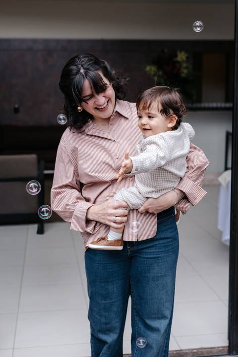 Mãe sorridente brincando com o bebê enquanto tentam pegar bolinhas de sabão no ar.'