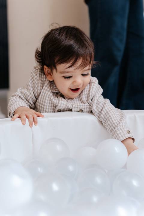Aniversariante sorrindo e se debruçando para brincar na piscina de bolinhas brancas.'