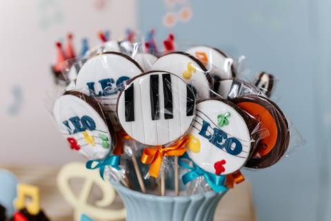 Detalhe dos pirulitos de biscoito decorados com o tema de notas musicais e teclado de piano.'