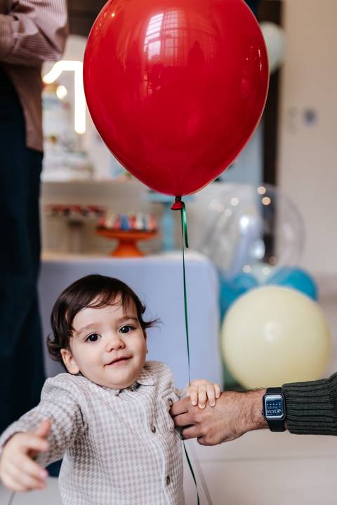 Aniversariante olhando para cima curioso com um balão vermelho ao seu lado.'