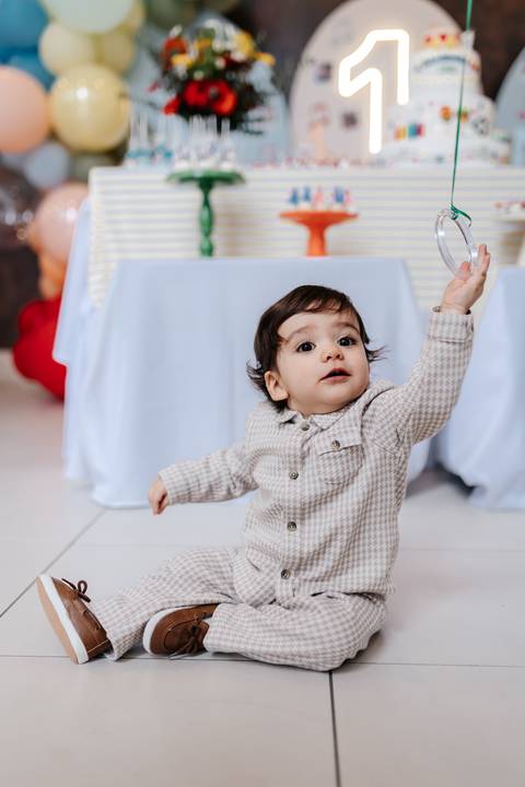 Aniversariante sentado no chão levantando a mãozinha para pegar algo no alto.'