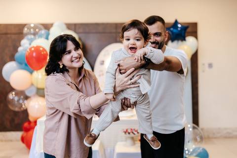 Pais brincando de erguer o aniversariante juntos no ar em momento de pura diversão.'