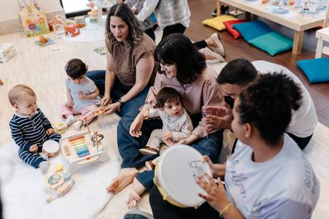 Grupo de mães e crianças sentadas no chão interagindo em uma roda de musicalização com pandeiro.'