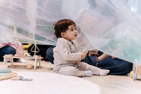 Bebê sentado no chão observando curioso por baixo de um tecido leve e transparente.'