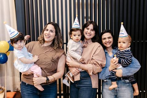 Mães sorridentes posando juntas com seus bebês usando chapéus de festa coloridos.'