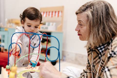 Bebê brincando com um aramado pedagógico sob o olhar atento de uma convidada.'