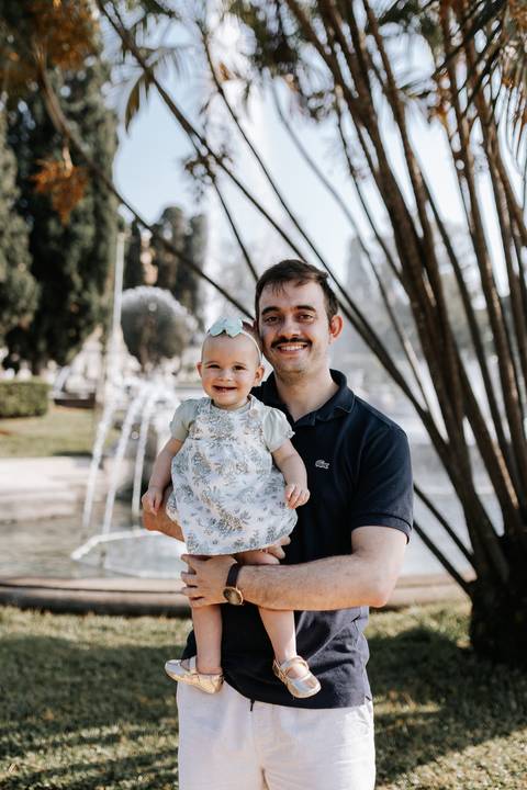 Pai sorridente segurando a filha no colo ao lado da fonte do parque.'