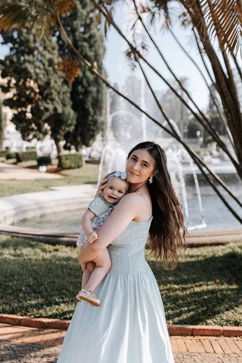 Mãe abraçando a bebê sorridente no colo com a fonte do parque ao fundo.'