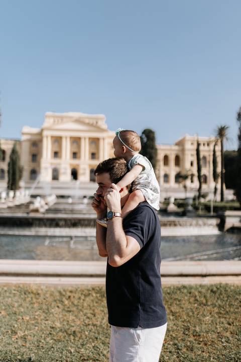 Pai carregando a bebê nos ombros enquanto caminham perto do espelho d'água do museu.'