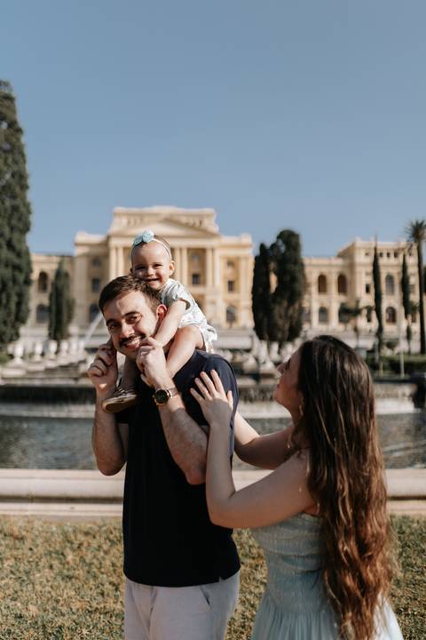 Pai com a filha nos ombros e mãe ao lado sorrindo com o Museu do Ipiranga ao fundo.'