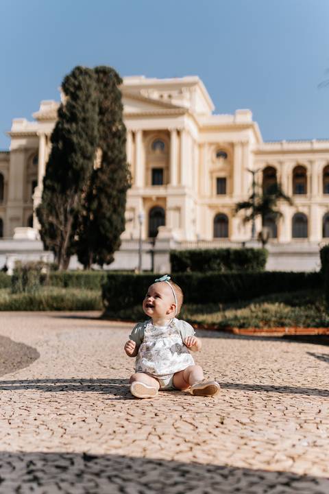 Bebê sentada no chão da praça com a imponente fachada do Museu do Ipiranga ao fundo.'
