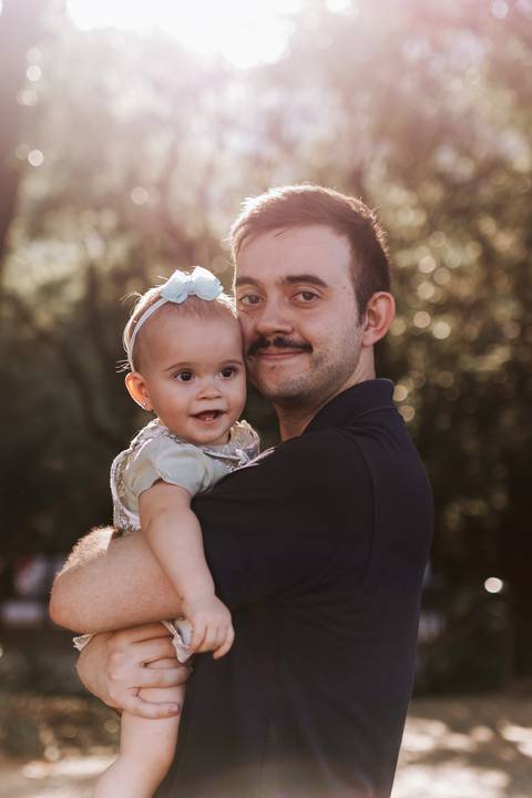 Pai sorrindo e segurando a bebê no colo entre as árvores do parque.'