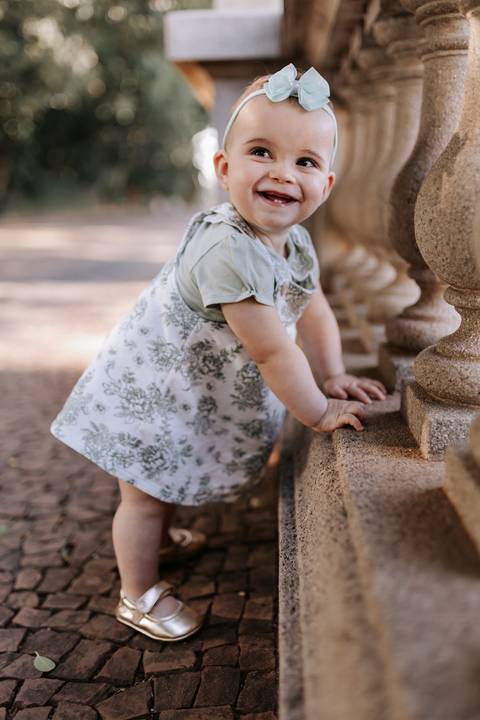 Bebê de vestido estampado apoiada na balaustrada de pedra explorando o espaço.'