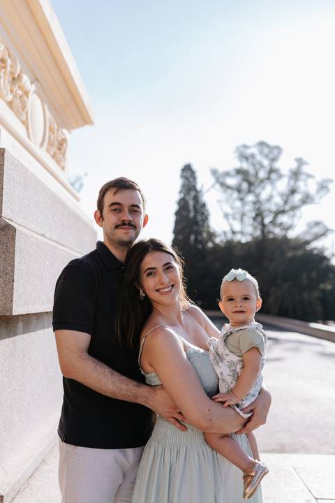 Casal e filha sorrindo para a câmera com a mureta de pedra e o parque ao fundo.'