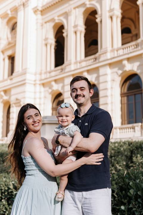 Retrato do casal sorridente com a bebê no colo em frente à fachada histórica.'