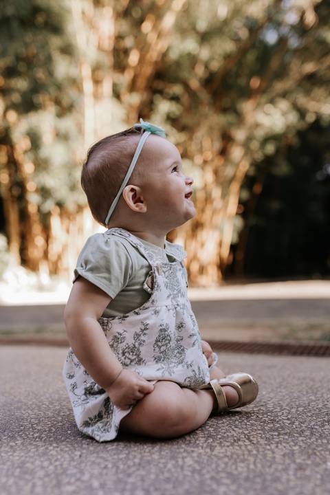 Bebê em pé no parque olhando para cima em meio à vegetação ao fundo.'