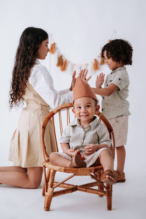 Bebê com coroa de pano na cadeira recebendo o carinho dos irmãos ao seu lado.'