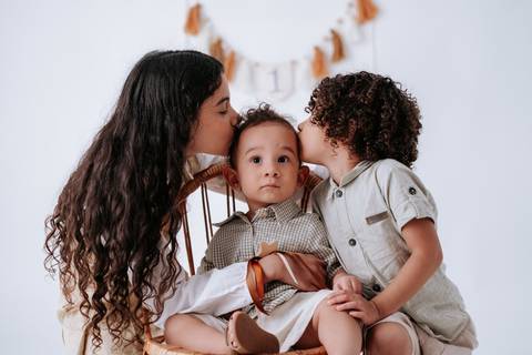 Irmãos mais velhos dando beijos carinhosos nas bochechas do bebê ao mesmo tempo.'