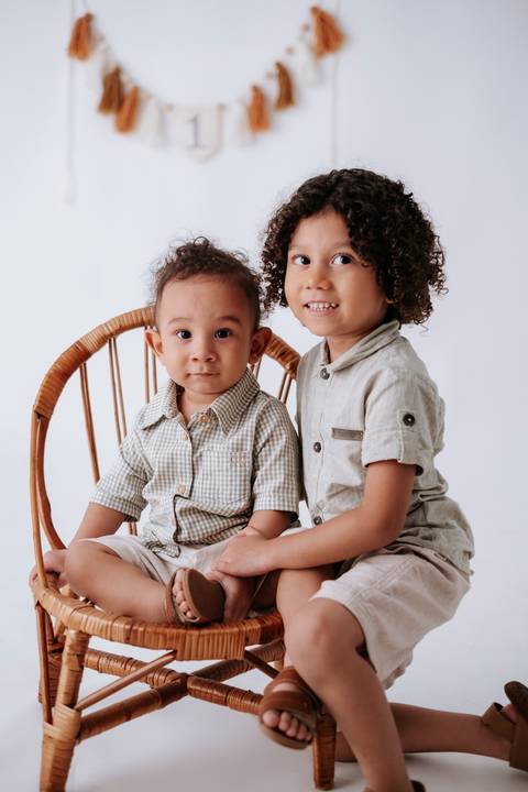 Bebê e o irmão mais velho sentados juntos na cadeira de palha sorrindo para a câmera.'