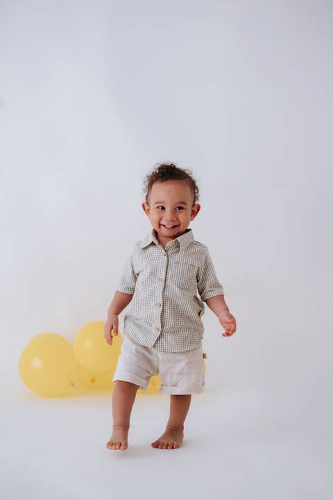 Bebê em pé no estúdio sorrindo com balões amarelos ao fundo.'