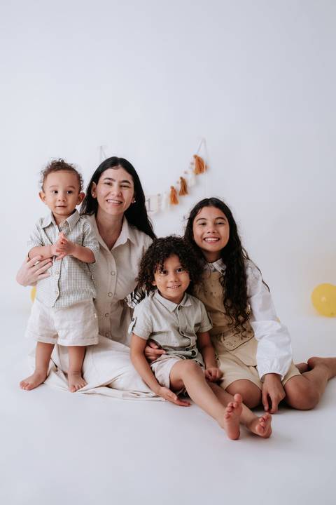 Mãe sorridente abraçada aos três filhos com o bebê em seu colo.'