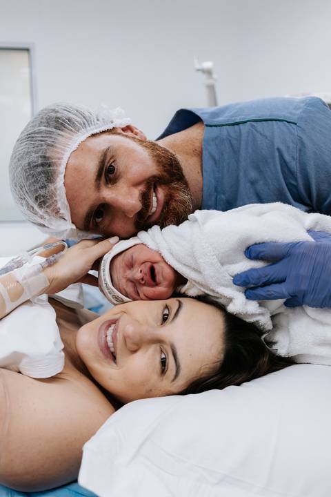 Retrato do casal sorrindo com o bebê enrolado no centro.'