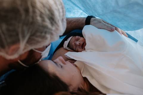 Bebê no contato pele a pele no colo da mãe com o pai ao lado.'